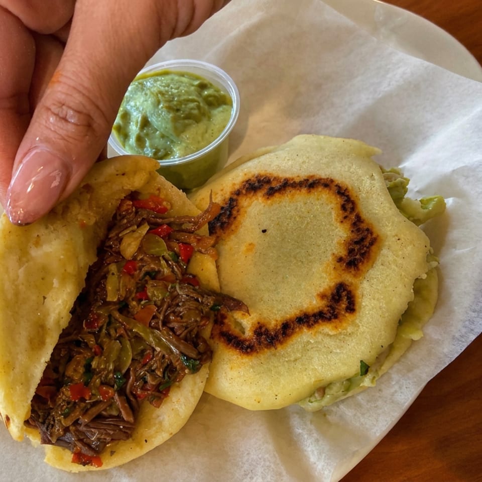 Open stuffed arepa with shredded beef, peppers, and onions beside a grilled arepa and cup of green sauce.